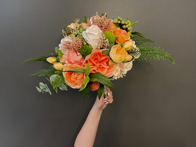 This radiant bouquet pairs deep green foliage with bight warm-toned flowers. Featuring peach peonies, orange roses, green and pink hypericum berries, cream hydrangea and miniature peaches. Accented with eucalyptus, ferns, cascading white berries and finished with a cream and brown gingham ribbon, secured with pearl pins.