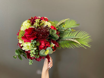 This bouquet offers a charcuterie of varietals. It features light green and dark scarlet hydrangeas, sage chrysanthemum, scarlet cone flowers, croton leaves, and rich burgundy roses, tied together with burgundy satin ribbon and secured with white pearl pins.
