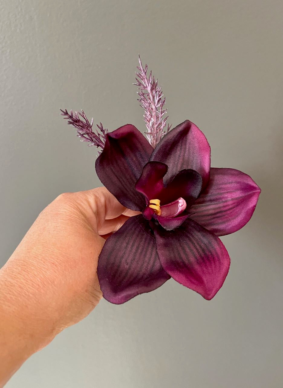 Boutonniere from our Black Cherry Collection. Crafted with a single deep, rich eggplant orchid and two sprigs of wild wispy lavender, it is artfully wrapped in a luxuriant deep plum satin.