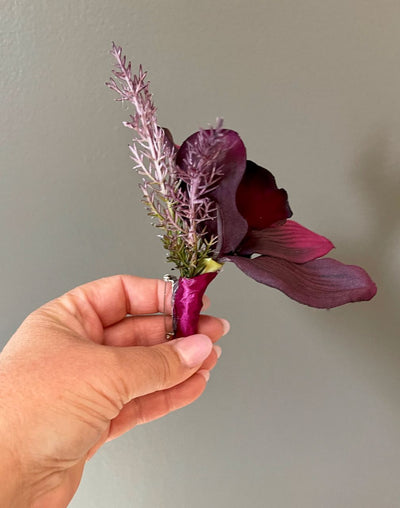 Boutonniere from our Black Cherry Collection. Crafted with a single deep, rich eggplant orchid and two sprigs of wild wispy lavender, it is artfully wrapped in a luxuriant deep plum satin.