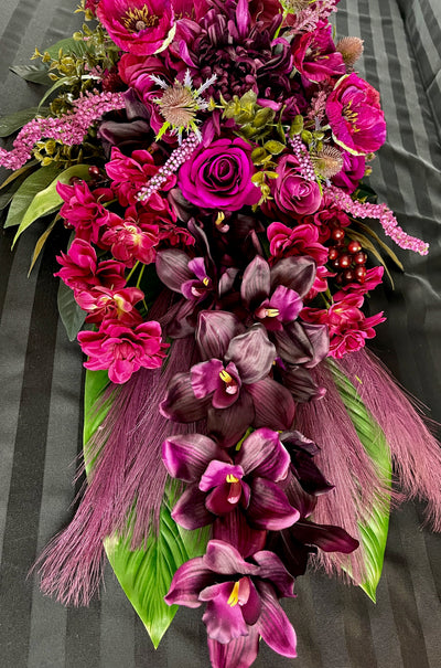 Introducing our Black Cherry Spray, a stunning blend of deep, rich hues and textures. Adorned with dahlias, roses, poppies, and hydrangea in shades of burgundy and fuchsia, complemented by thistle, eucalyptus, and spiral amaranth. The base is adorned with dark green palm leaves, purple feathers, and luxurious orchids. Experience the luxury and exclusivity of this four-foot masterpiece.