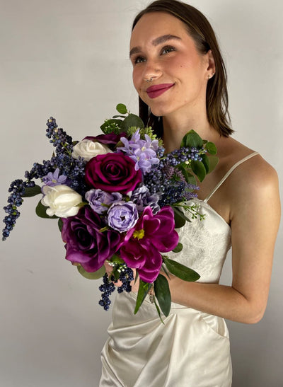 This vibrant jewel toned bridesmaid bouquet features a gradient of rich violet and fuchsia roses, white chrysanthemum, pale purple dahlia, amethyst hydrangea, thistle, lavender, and green succulent sprigs.This bouquet rents for $59.00 for five days.