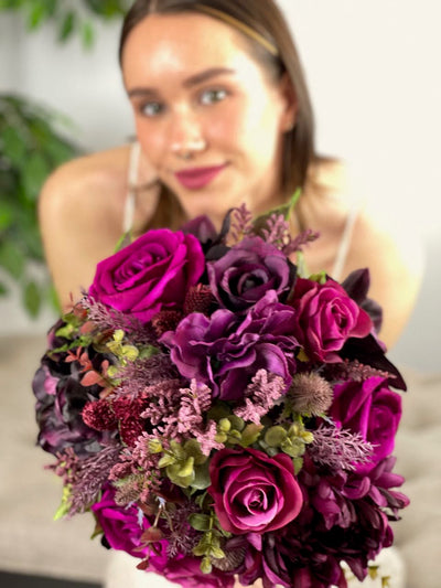 This stunning bridal bouquet merges the freshness of magenta roses with the regal presence of purple hydrangea and black orchids, all elegantly offset by the luxuriant shades of plum, deep green eucalyptus sprigs, and burgundy thistle. Expertly crafted into a hand-tied bouquet and finished with sumptuous amethyst satin ribbon, this statement piece is the perfect complement for your bridal ensemble. Rent for Five days for $88.00