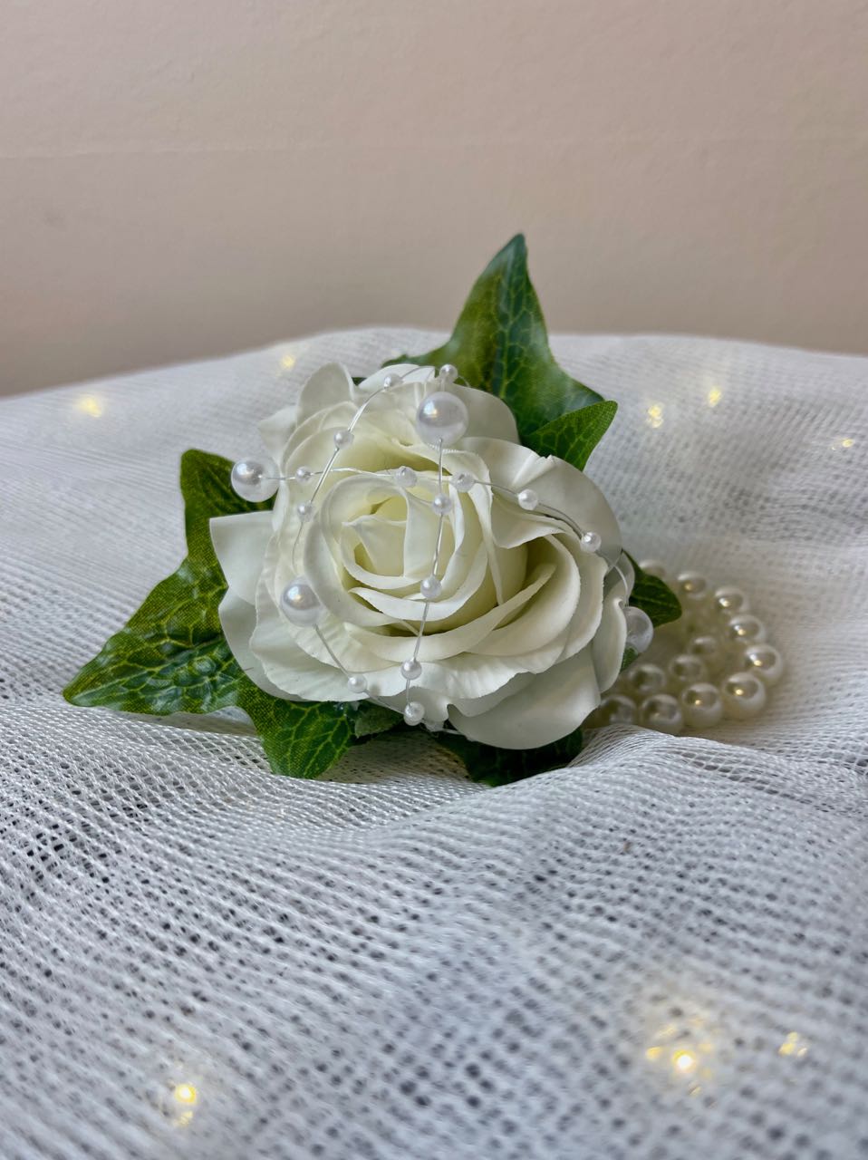 This beautiful wrist corsage features a single white rose nestled in a bed of ivy leaves, securely fastened to a pearl bracelet. A string of various sized pearls crisscrossing the rose adds visual interest, making this an elegant and sophisticated accessory for any special occasion