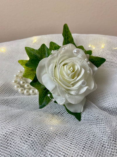 This beautiful wrist corsage features a single white rose nestled in a bed of ivy leaves, securely fastened to a pearl bracelet. A string of various sized pearls crisscrossing the rose adds visual interest, making this an elegant and sophisticated accessory for any special occasion