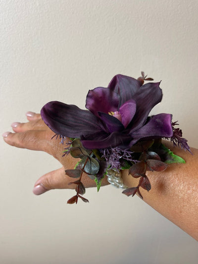 This exquisite Wrist Corsage in Black Cherry offers a sophisticated yet unique statement for special occasions. Crafted with a single deep eggplant colour orchid and sprigs of wild lavender, burgundy, and dark green eucalyptus, it is artfully arranged on a pearl bracelet and exudes understated elegance.