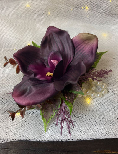 This exquisite Wrist Corsage in Black Cherry offers a sophisticated yet unique statement for special occasions. Crafted with a single deep eggplant colour orchid and sprigs of wild lavender, burgundy, and dark green eucalyptus, it is artfully arranged on a pearl bracelet and exudes understated elegance.
