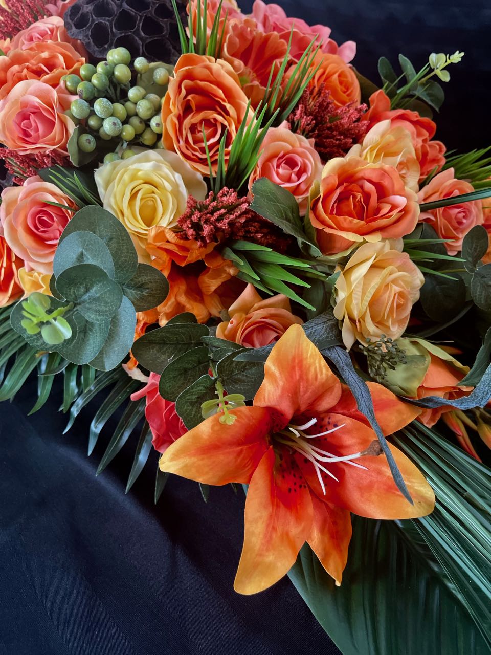 This vivid floral arrangement, a tribute to the fiery hues of a summer sunset, juxtaposes the lushness of palm leaves, eucalyptus sprigs, and tall grasses with the vividness of tiger lillies, green hypercium berries, and an array of roses in sundry shades of orange and brown-hued honeycombs. This arrangement is four feet long and 16 inches wide.