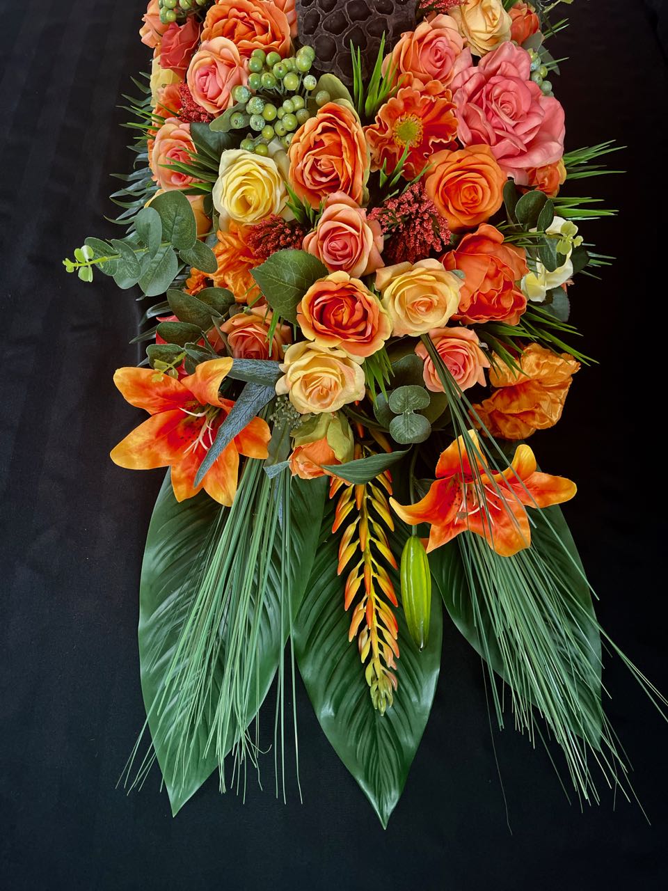 This vivid floral arrangement, a tribute to the fiery hues of a summer sunset, juxtaposes the lushness of palm leaves, eucalyptus sprigs, and tall grasses with the vividness of tiger lillies, green hypercium berries, and an array of roses in sundry shades of orange and brown-hued honeycombs. This arrangement is four feet long and 16 inches wide.