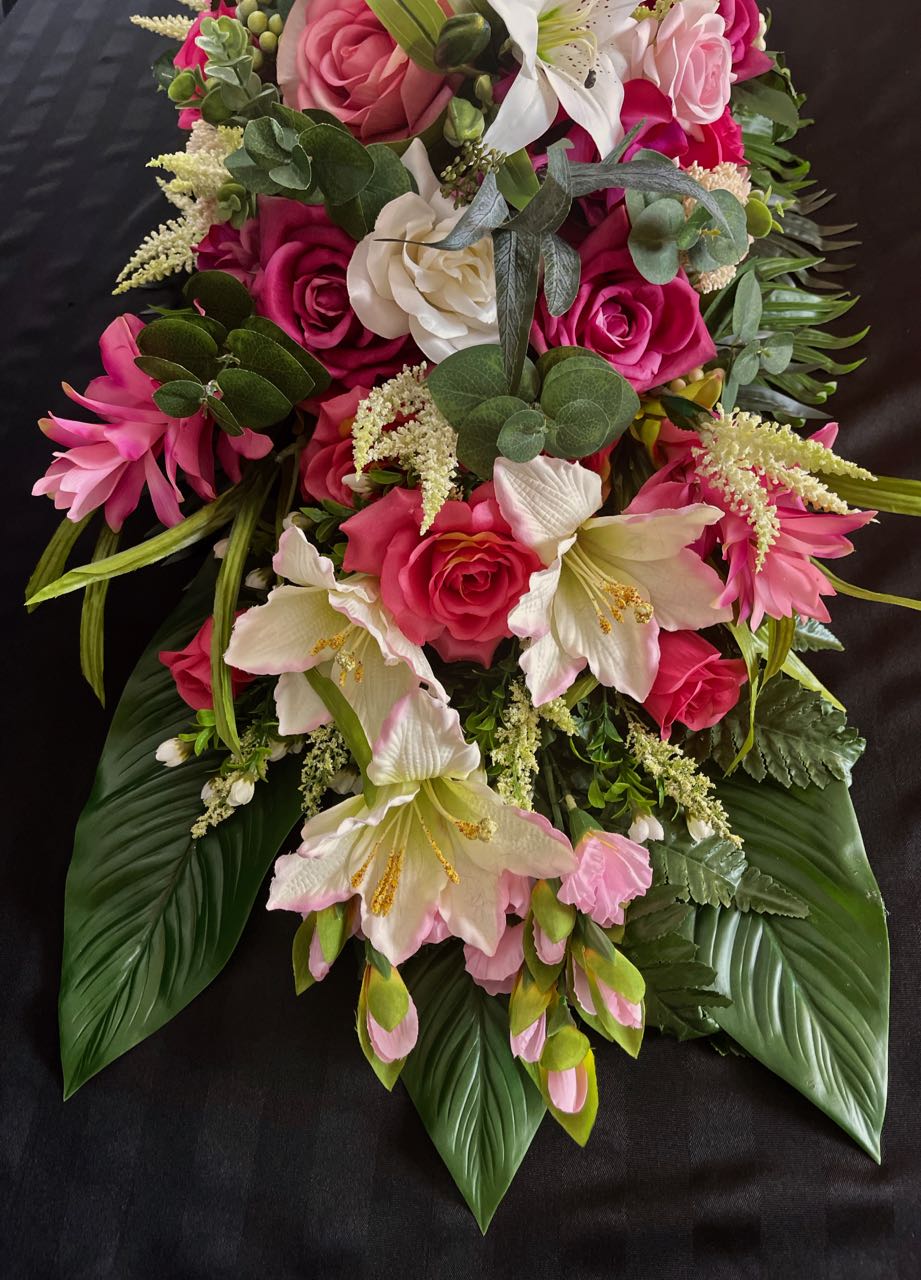 This vivid, lush tapestry of color and texture combines cream dahlias, huge fuschia roses, and white lilies kissed with pink edges. To add even more visual interest this design is accented with green hypericum berries, eucalyptus sprigs, and cream astilbe playfully poking out between flowers. The base of this four foot long and 16 inch wide creation is dark green shiny palm leaves that gives this creation a tropical feel.