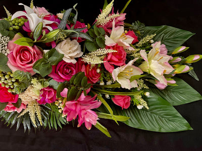 This vivid, lush tapestry of color and texture combines cream dahlias, huge fuschia roses, and white lilies kissed with pink edges. To add even more visual interest this design is accented with green hypericum berries, eucalyptus sprigs, and cream astilbe playfully poking out between flowers. The base of this four foot long and 16 inch wide creation is dark green shiny palm leaves that gives this creation a tropical feel.