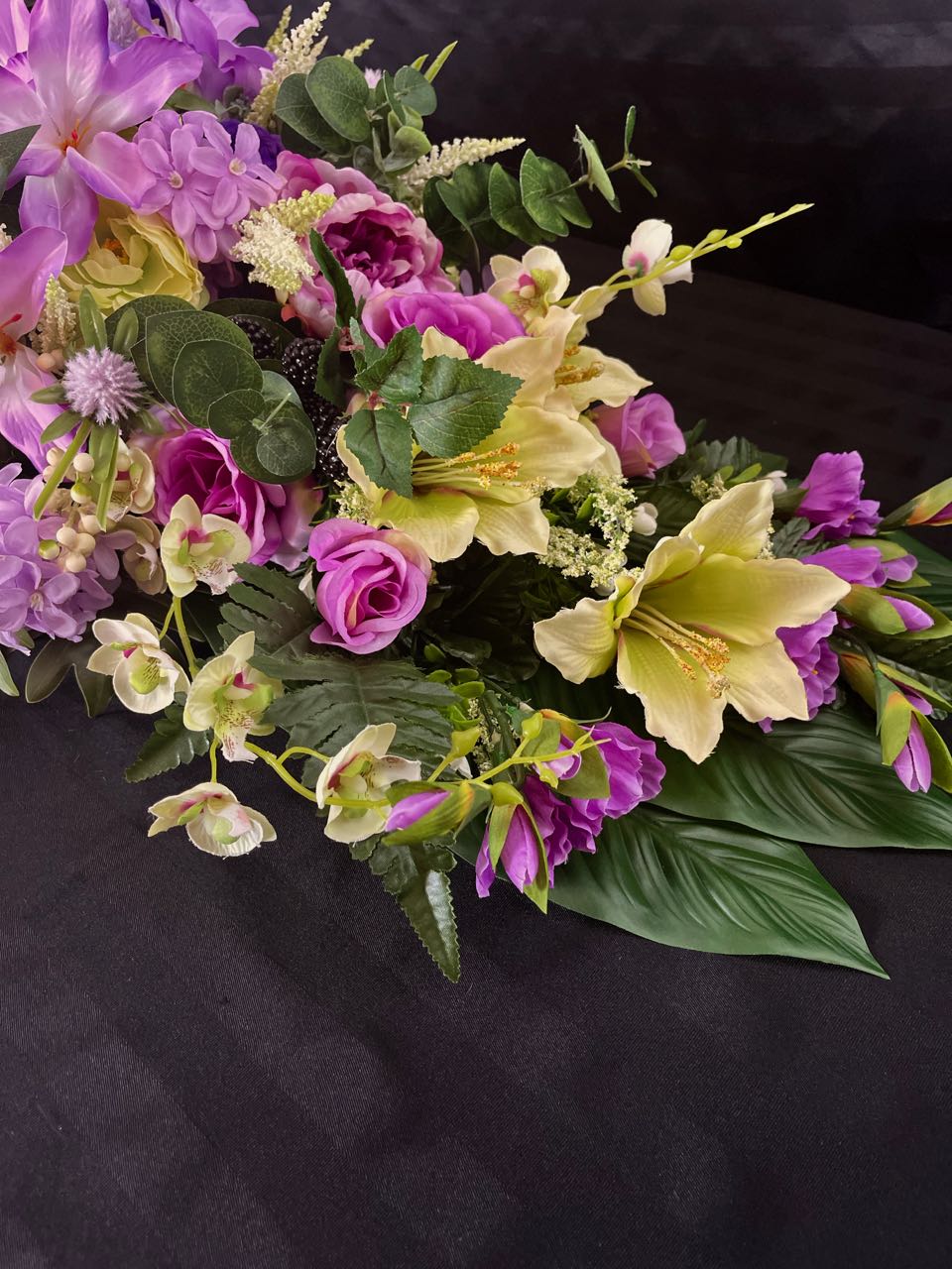 Indulge in the lushness of this vibrant floral arrangement. Deep purple and delicate mauve roses are artfully paired with subtly light lime green lilacs. Majestic purple peonies, pom pom dahlias, orchids, and lilies coalesce to create a stunning vision. This design measures four feet long and 16 inches wide, elevated upon glossy hunter green palm leaves.