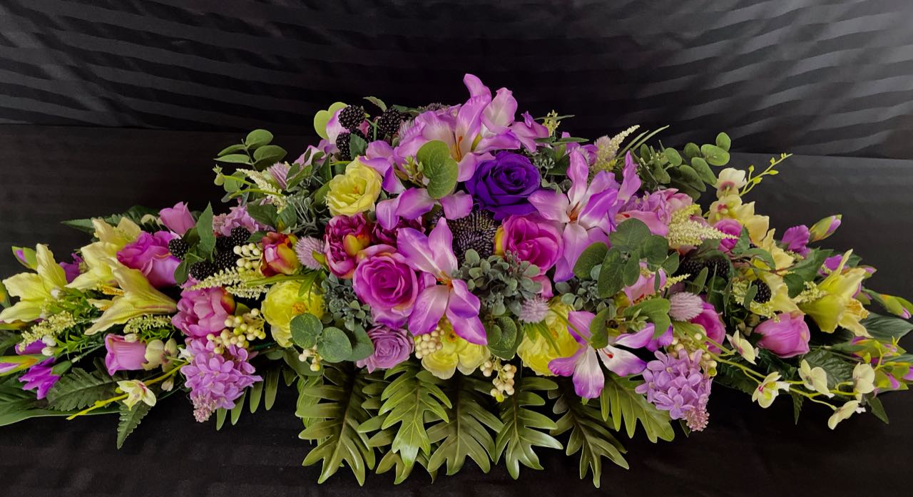 Indulge in the lushness of this vibrant floral arrangement. Deep purple and delicate mauve roses are artfully paired with subtly light lime green lilacs. Majestic purple peonies, pom pom dahlias, orchids, and lilies coalesce to create a stunning vision. This design measures four feet long and 16 inches wide, elevated upon glossy hunter green palm leave