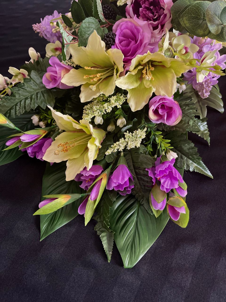 Indulge in the lushness of this vibrant floral arrangement. Deep purple and delicate mauve roses are artfully paired with subtly light lime green lilacs. Majestic purple peonies, pom pom dahlias, orchids, and lilies coalesce to create a stunning vision. Enjoy the unexpected accent of blackberries popping out of the flowers to add visual interest. This design measures four feet long and 16 inches wide, elevated upon glossy hunter green palm leaves.