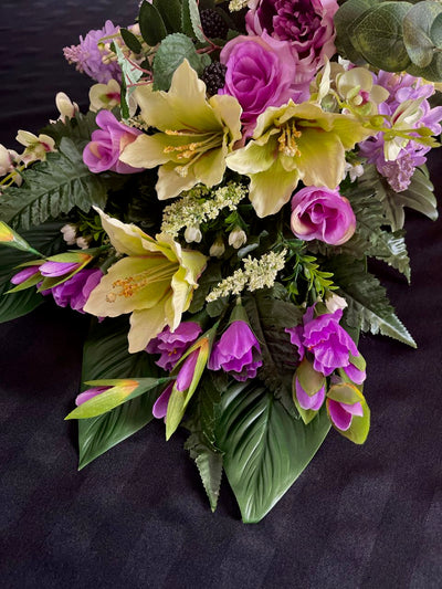 Indulge in the lushness of this vibrant floral arrangement. Deep purple and delicate mauve roses are artfully paired with subtly light lime green lilacs. Majestic purple peonies, pom pom dahlias, orchids, and lilies coalesce to create a stunning vision. Enjoy the unexpected accent of blackberries popping out of the flowers to add visual interest. This design measures four feet long and 16 inches wide, elevated upon glossy hunter green palm leaves.