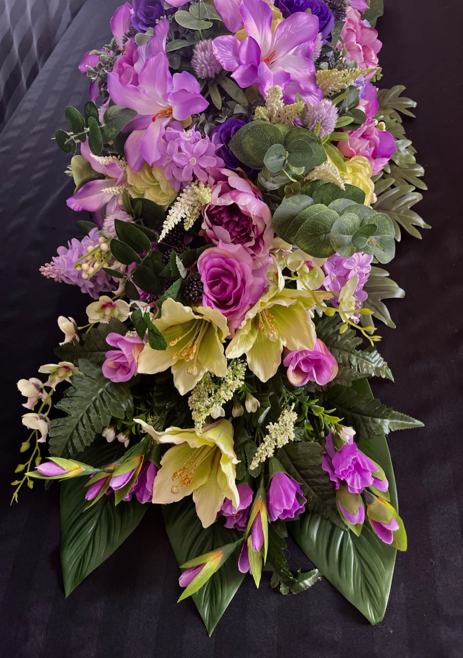 Indulge in the lushness of this vibrant floral arrangement. Deep purple and delicate mauve roses are artfully paired with subtly light lime green lilacs. Majestic purple peonies, pom pom dahlias, orchids, and lilies coalesce to create a stunning vision. This design measures four feet long and 16 inches wide, elevated upon glossy hunter green palm leaves.
