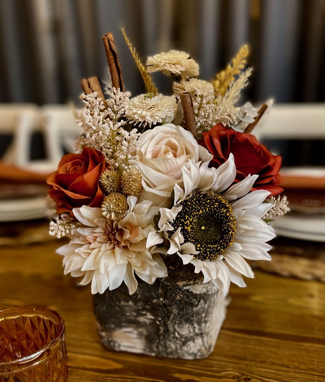 This centrepiece features rust and cream roses combined with pampas, eucalyptus, and for a fun twist, cinnamon sticks, all nestled in a birch vase. The centrepiece is approx 8 inches wide by 12 inches tall. This earthy concoction would not look out of place on a cocktail table,  however it has enough presence and height to be used on a 8 foot round tale surrounded by candle votives.