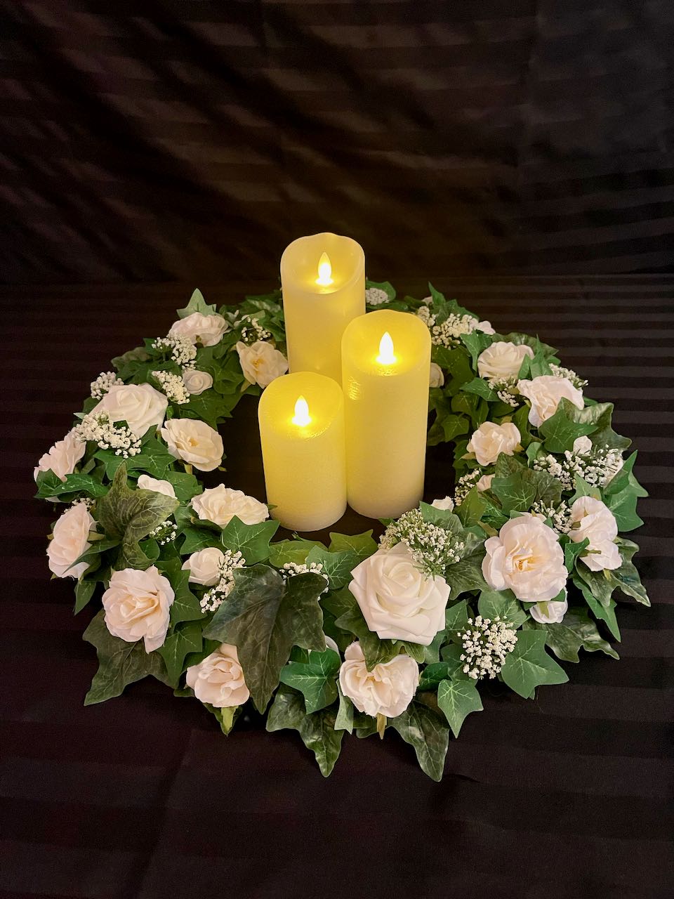 Dark green ivy with various sizes of white roses interspersed between the leaves create this simple yet elegant centrepiece. The ring can be used alone but we love the look of adding in cream pillar candles. Select the number of LED pillar candles you would like to complete this look. The outer circumference of this wreath is 21 inches and in inner circumference is 10 inches. Pillar candles to be rented separately. Each pillar candle is an additional $3.00 to $4.00 to rent.