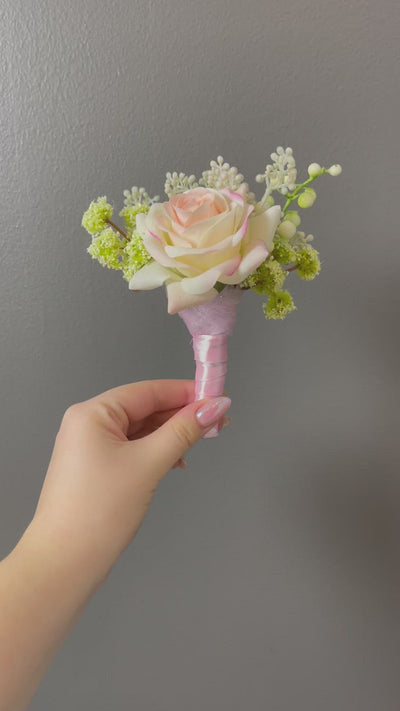 This delicate 6" boutonniere features a cream and light pink rose, and various green foliage, secured with a soft pink satin ribbon. 