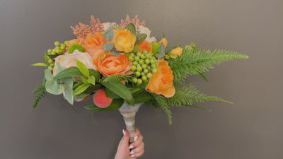 This radiant bouquet pairs deep green foliage with bight warm-toned flowers. Featuring peach peonies, orange roses, green and pink hypericum berries, cream hydrangea and miniature peaches. Accented with eucalyptus, ferns, cascading white berries and finished with a cream and brown gingham ribbon, secured with pearl pins.