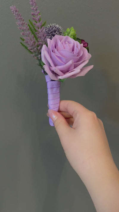 This 6" boutonniere features a light purple rose, sprigs of lavender, thistle, eggplant rose, and evergreen foliage detail, finished with a light purple ribbon.