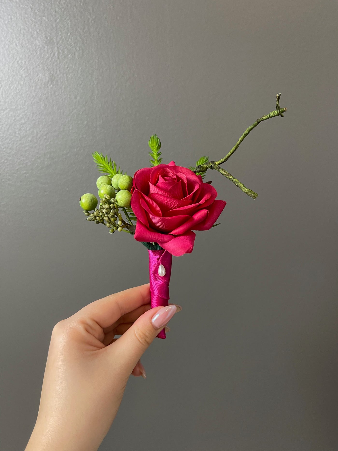 This 6" sophisticated boutonniere features a scarlet rose, green hypericum berries, and assorted foliage. It is secured with satin plum ribbon and a pearl teardrop pin.