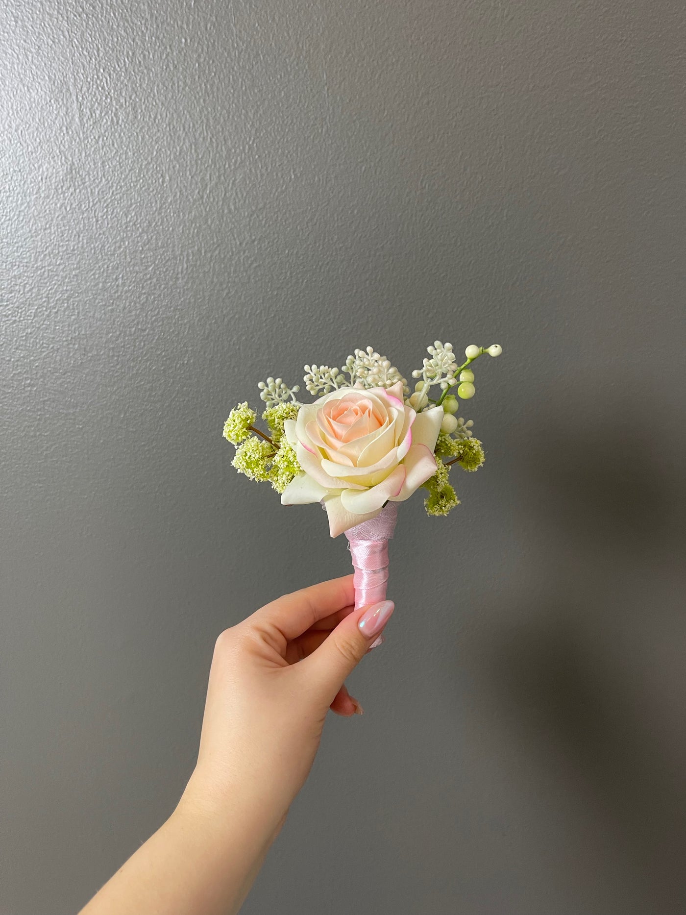 This delicate 6" boutonniere features a cream and light pink rose, and various green foliage, secured with a soft pink satin ribbon. 