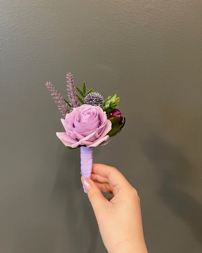 This 6" boutonniere features a light purple rose, sprigs of lavender, thistle, eggplant rose, and evergreen foliage detail, finished in a light purple ribbon.