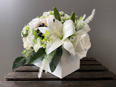 This delicate white and cream arrangement features a variety of roses, lilies, hydrangeas, dahlia, peonies, lilacs, baby’s breath perched on a bed of ferns and artfully arranged in a white box with pearl detail (L8”xW4”xH4”).