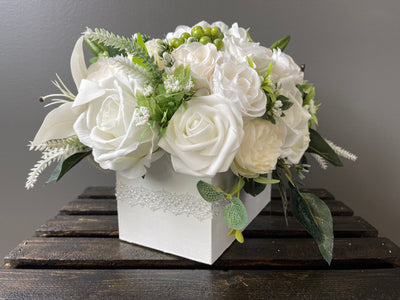 This delicate white and cream arrangement features a variety of roses, lilies, hydrangeas, dahlia, peonies, lilacs, baby’s breath perched on a bed of ferns and artfully arranged in a white box with pearl detail (L8”xW4”xH4”).