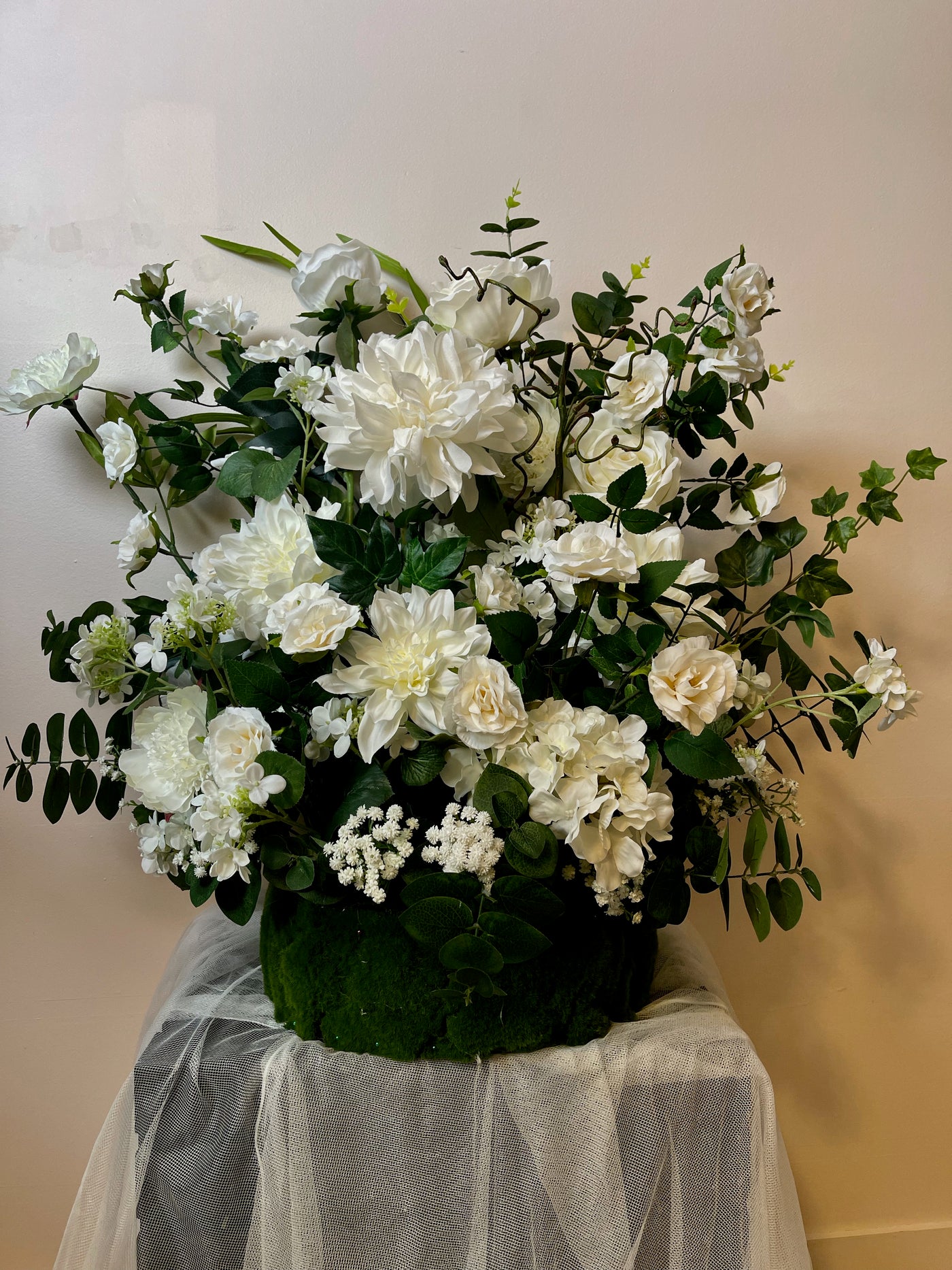 Crisp white dahlias, roses and baby's breath enveloped in dark green eucalyptus  are combined to create this dramatic 2.5 x 3 foot wedding floral arrangement. This was designed to be used at the base of a floral arch but it can also be used at the base of a sweetheart head table, or on either side of the alter at a church. The options are endless.