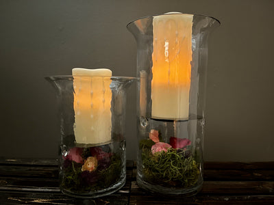 A set of two thick glass vases (12" and 9") that include realistic LED dripping wax candles. Under a black plate in the vase is an arrangement of multi coloured bud roses on a floor of three different types of moss