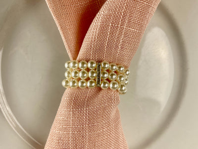 Elegant napkin ring comprised of three stands of pearls with a silver clasp studded with diamonds holding them together. Shown on a pink linen napkin.Rent for $.25