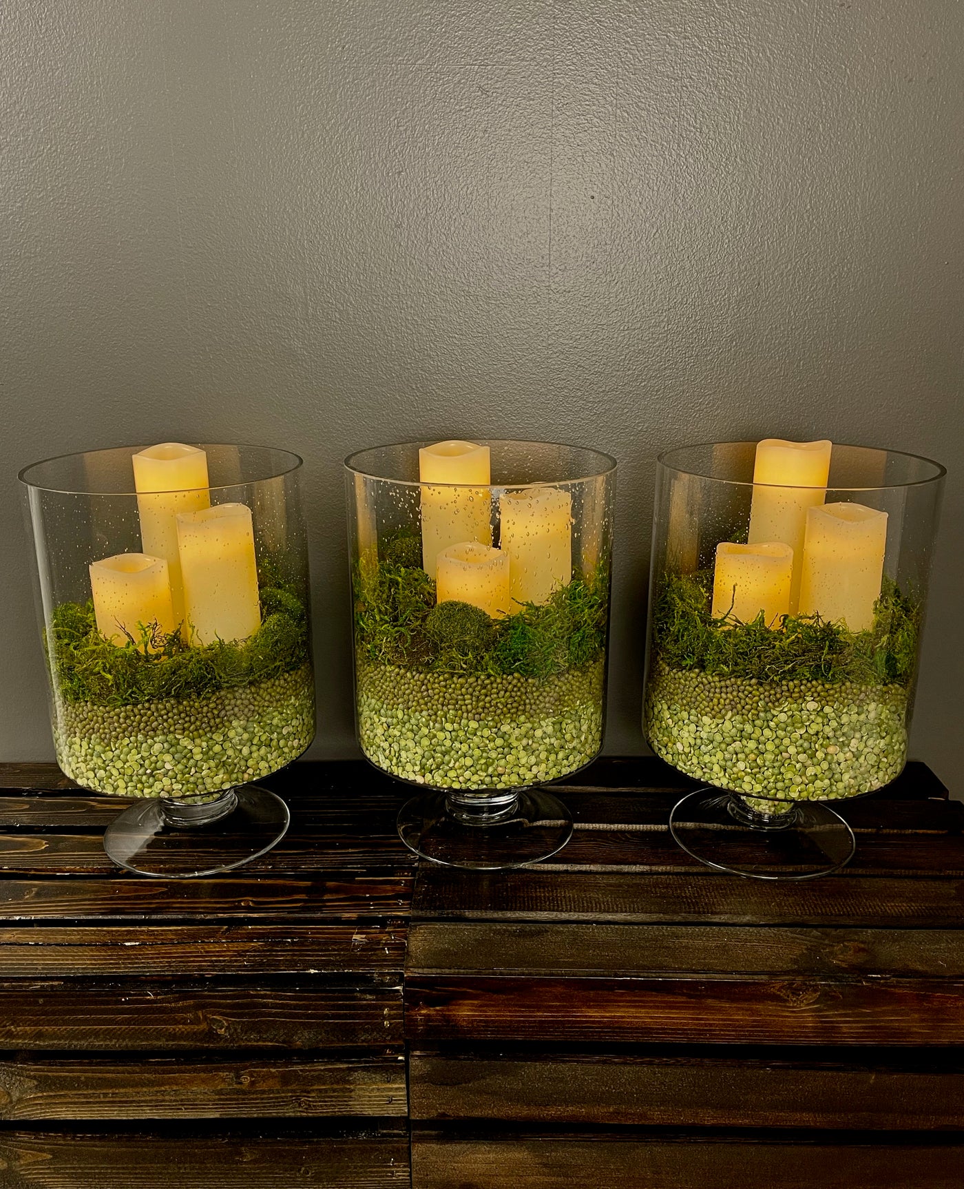 Set of three thick glass 12" pedestal vases, each holding three remote controlled LED Ivory candles nestled in a deep rich moss base
