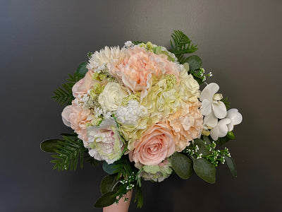 This soft pastel bouquet is the perfect whisper of elegance featuring pale pink roses, blush dahlia, delicate white peonies, pale pink cabbage rose, white sweet peas, hydrangea, baby’s breath, and accented with miniature viburnum. Three white orchids cascade down asymmetrically, all sitting atop a bed of seeded eucalyptus and ferns. Bound delicately with baby pink satin ribbon.