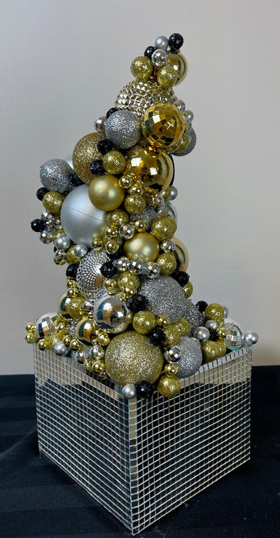 Silver, black and gold balls of all shapes and textures are stacked on top of each other to create this futuristic Skyscaper. This fun structure is busting out of a mirrored Disco ball box. At 12 inches tall and five inches wide this is a perfect size for a stand-up cocktail table. Each piece is unique with some having more gold or silver than others.