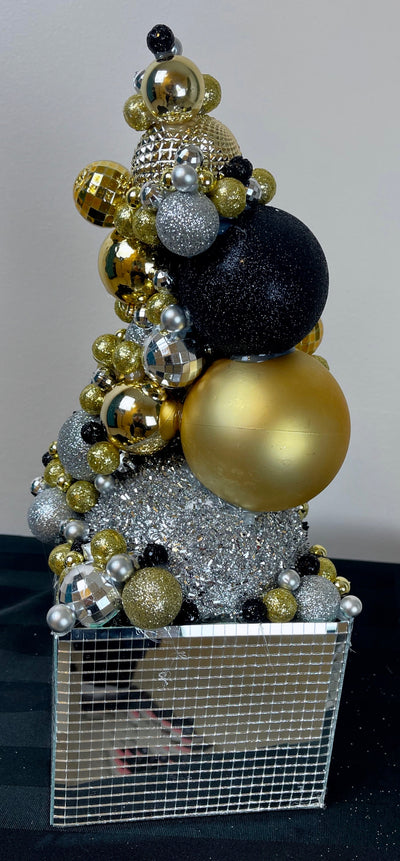 Silver, black and gold balls of all shapes and textures are stacked on top of each other to create this futuristic Skyscaper. This fun structure is busting out of a mirrored Disco ball box. At 12 inches tall and five inches wide this is a perfect size for a stand-up cocktail table. Each piece is unique with some having more gold or silver than others.
