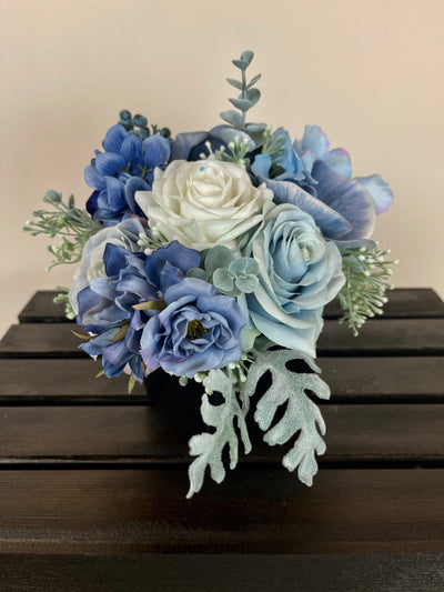 This sophisticated medley of navy, light blue, and white roses, hydrangeas, orchids, and blueberries is accented by eucalyptus and dusty miller foliage to craft a gorgeous centrepiece for a raised cocktail table. Measuring 4" x 4", the florals are cascading our of a square glossy black vase.