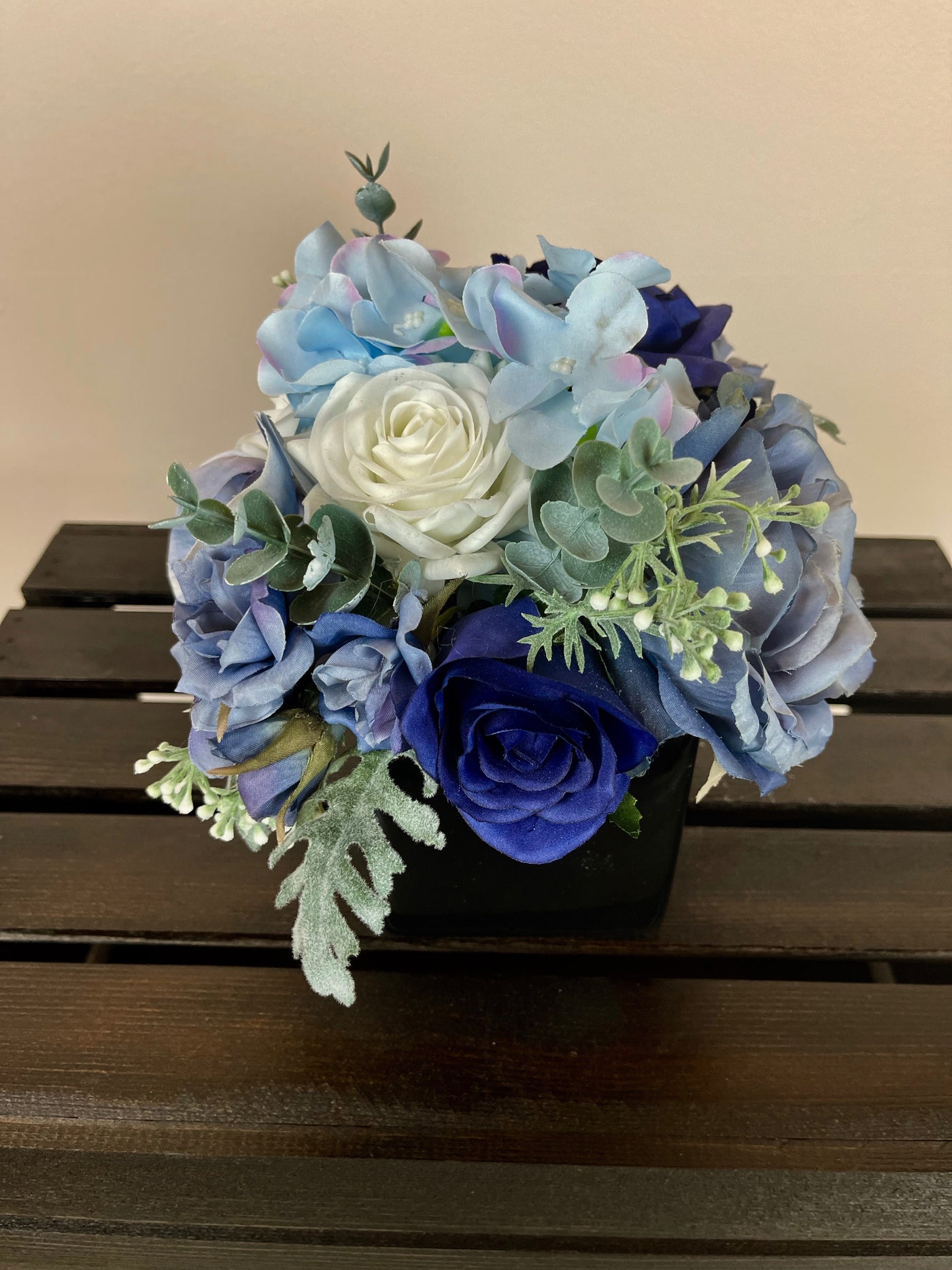 This sophisticated medley of navy, light blue, and white roses, hydrangeas, orchids, and blueberries is accented by eucalyptus and dusty miller foliage to craft a gorgeous centrepiece for a raised cocktail table. Measuring 4" x 4", the florals are cascading our of a square glossy black vase.