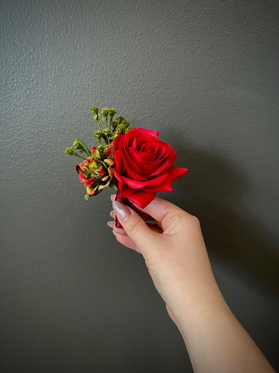 This stunning five inch boutonniere has a single dark red rose accompanied by a red and green variegated ranunculus nestled in a cloud of rich green foliage.