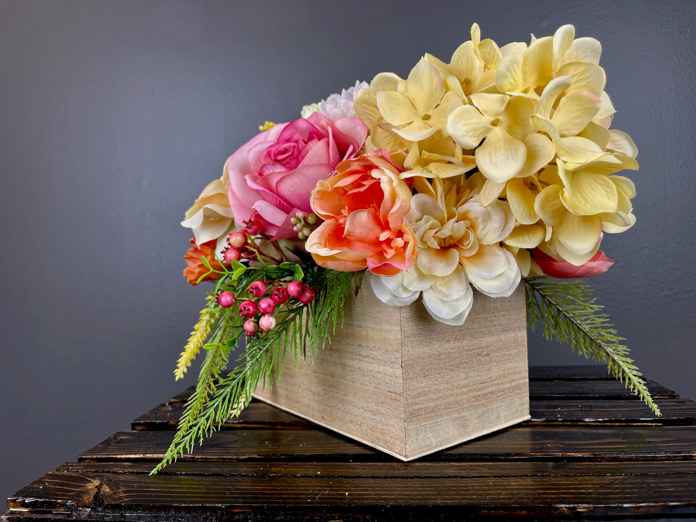 Invite the colorful array of a summer sunset  inside with this bright centrepiece. Large pink and yellow peony blossoms open gracefully next to custard hydrangeas, peach English roses, white globe dahlia, kicky craspedia, and yellow hypericum berries cascading alongside bright green fern. Placed in a lacquered pine box (L8”x W4”x H4”).