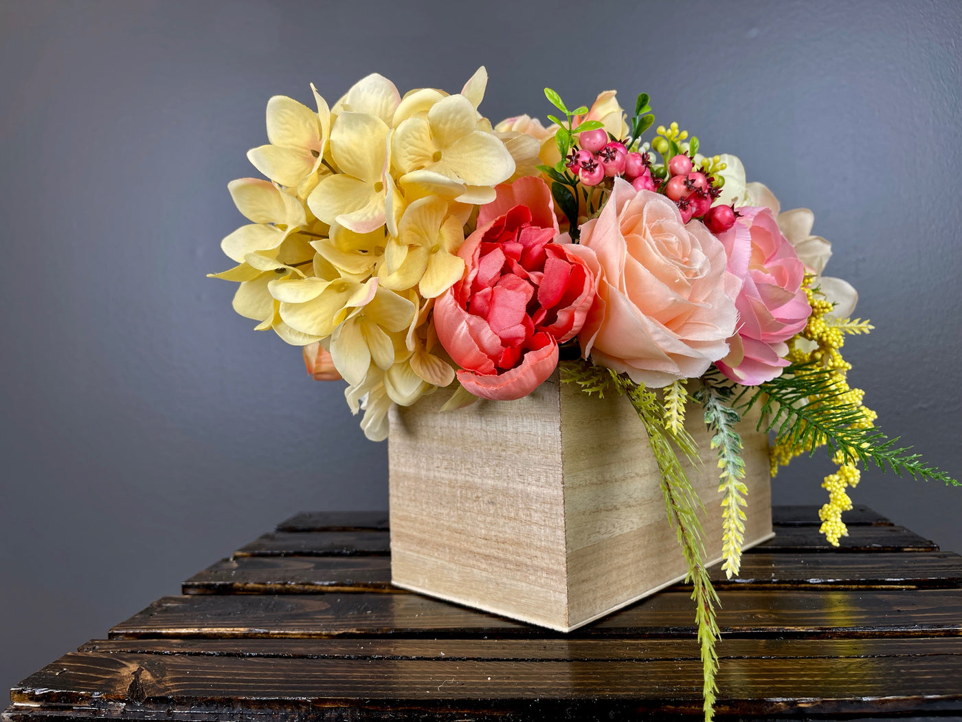 Invite the colorful array of a summer sunset  inside with this bright centrepiece. Large pink and yellow peony blossoms open gracefully next to custard hydrangeas, peach English roses, white globe dahlia, kicky craspedia, and yellow hypericum berries cascading alongside bright green fern. Placed in a lacquered pine box (L8”x W4”x H4”).