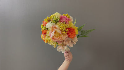 This warm summer bouquet features large pink and yellow peony blossoms which open gracefully next to custard hydrangeas, peach English roses, white globe dahlia, and yellow hypericum berries cascading alongside bright green fern. It is finished with light pink satin ribbon and secured with pearl teardrop pins.