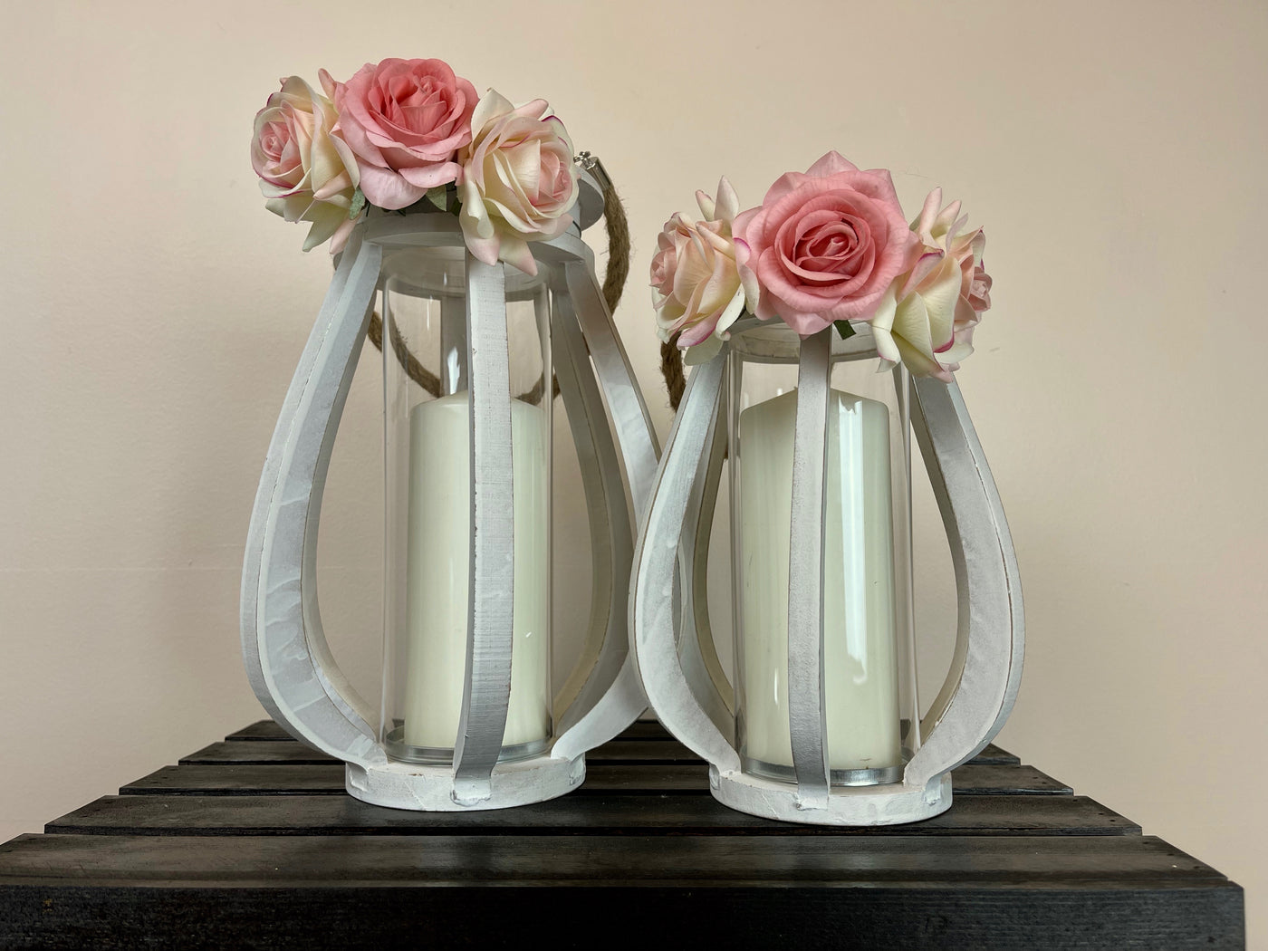 Two white hurricane lanterns of different sizes with a trio of roses attached to the top. 